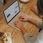 Person typing on laptop keyboard while researching spotlight on industry trends, working from a wooden table with food nearby, showcasing modern remote work lifestyle