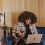 Person with curly hair sitting comfortably on a blue couch in a wood-paneled room, working on a laptop to master social media platforms for effective digital marketing